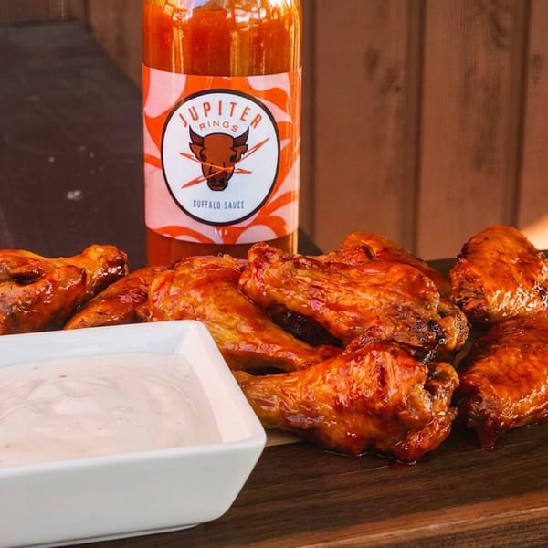 Glazed chicken wings, Jupiter Rings buffalo sauce bottle, and white dip on a wooden board.