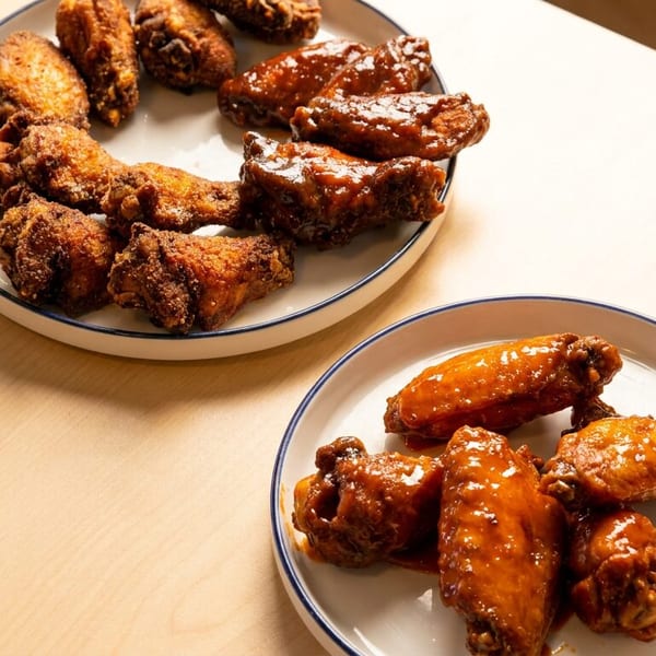Two plates of crispy and glossy sauced chicken wings on a light wooden table.